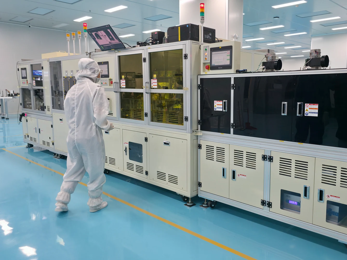 A worker in full cleanroom clothing stands beside enclosed automated machines on the blue floor of the production area.